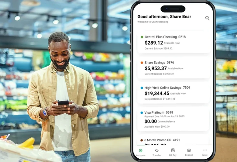 Person using a smartphone in a grocery store with a mobile banking app displayed on the screen showing account balances, savings, and credit card details.