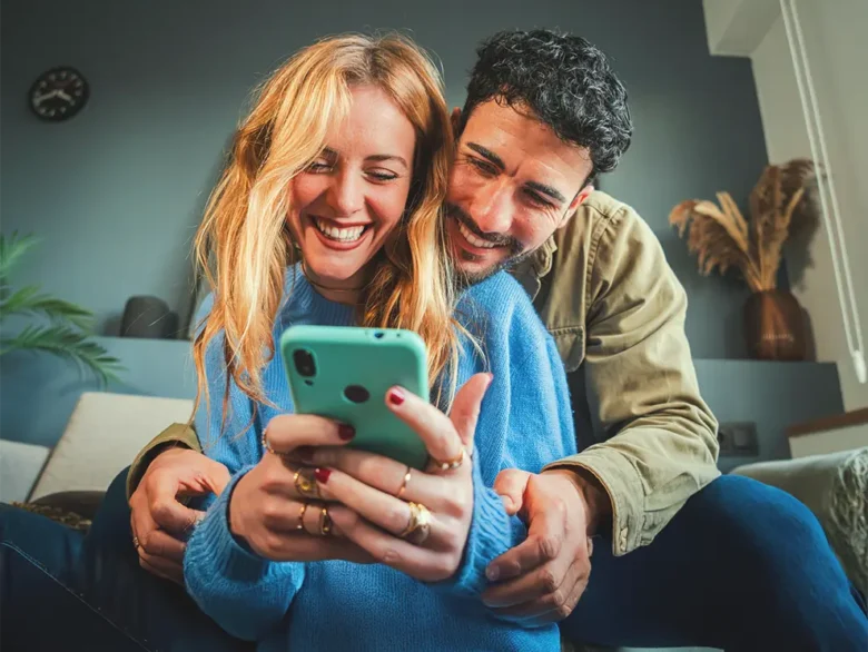 Couple on couch smiling at smartphone, cozy home setting.